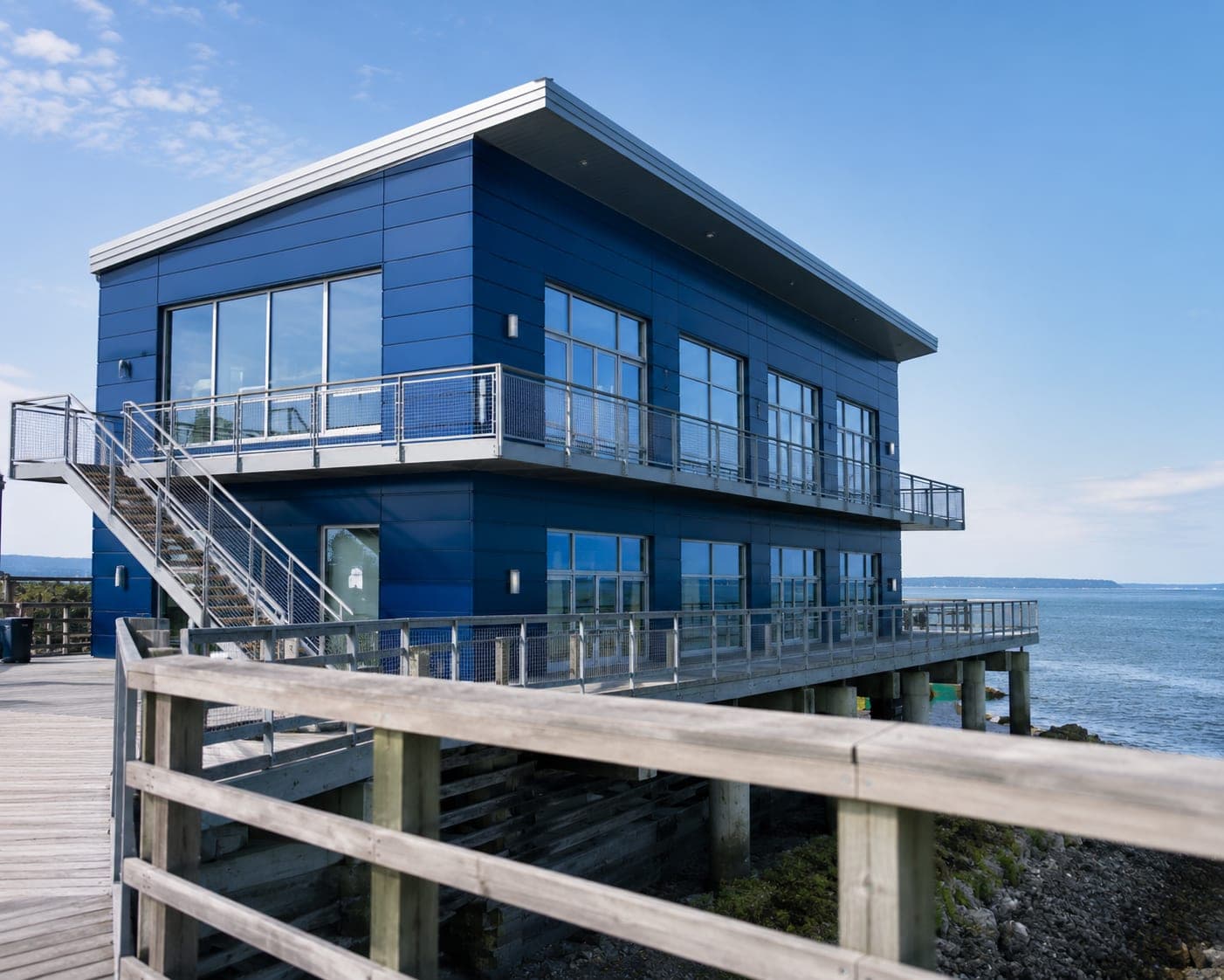 Two-story coastal building clad in deep marine-blue metallic aluminum panels along a waterfront boardwalk